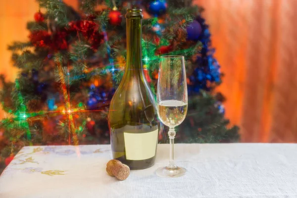Bottle and wine glass of white sparkling wine on a table on a blurred background of Christmas tree with festive lights