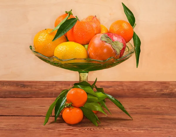 Different fresh fruits in the vintage green glass vase for fruits, small branch of tangerine tree with leaves and tangerines on the rustic table, top view in selective focus