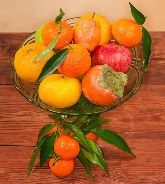 Different fresh fruits in the vintage green glass vase for fruits, small branch of tangerine tree with leaves and tangerines on the rustic table, top view in selective focus