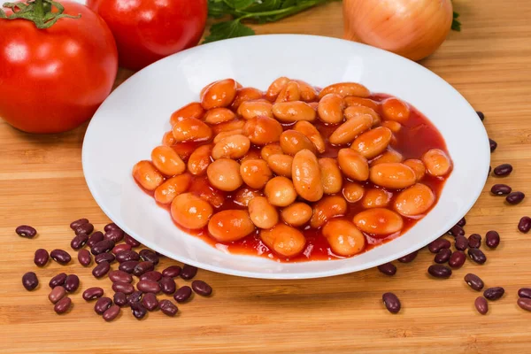 Boiled beans stewed with tomato sauce on a white dish against the some ingredients on a wooden surface