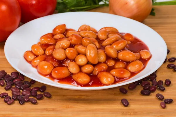 Boiled beans stewed with tomato sauce on a white dish on a wooden surface, close-up in selective focus