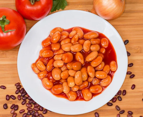 Boiled beans stewed with tomato sauce on a white dish on a wooden surface, top view