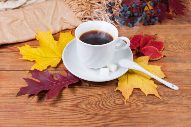 Beyaz bardakta kahve, çay tabağı ve kırsal bir masada küçük seramik kaşık, sonbahar yaprakları ve kadın atkısı.