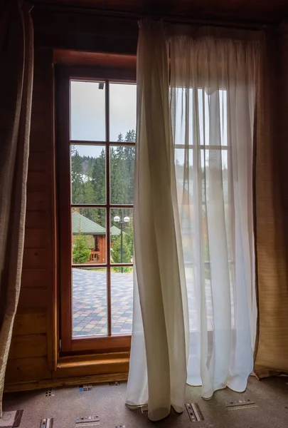 Large floor to ceiling window with thin wooden frames partly covered with curtains in a wooden house, inside view