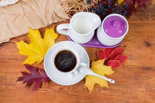 Coffee in white cup with saucer and small ceramic spoon, milk jug and sugar bowl on an old rustic table with autumn leaves and women\'s scarf, top view