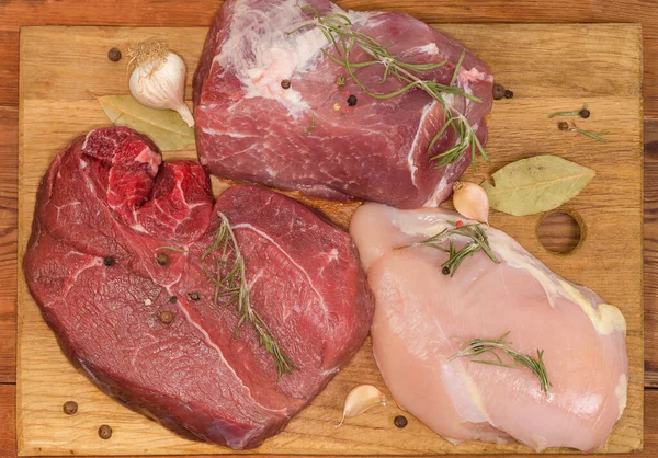様々な調理されていない骨のない肉の大きな部分 古い素朴なテーブルの上に木製のまな板の上に異なるスパイスで足の皮のない鶏の胸肉 豚肉と牛肉 トップビュー — ストック写真