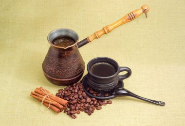 Siyah seramik fincanda sade Türk kahvesi ve klasik bakır kahve demliğinde, tarçın çubuklarında ve tekstil yüzeyine saçılmış kahve çekirdekleri.