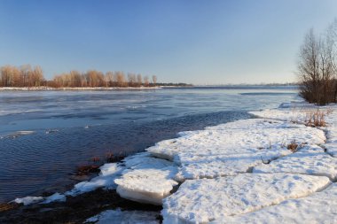 Geniş, düz nehir kısmen ince buzla kaplı, kıyıda kalın buzlar açık gökyüzüne karşı yüzüyor ve ilkbaharın başlarında karşı kıyıda ağaçlar var.
