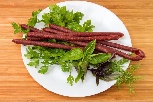 Long thin dry cured sausages in natural casing on a dish among the different fresh greens on a wooden surface, top view