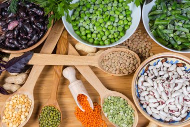 Farklı kaselerde çeşitli çiğ ve donmuş baklagiller ve ahşap kesme tahtasındaki tahta kaşıklar, üst kısmı yakın plan.