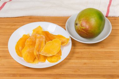Dried slices of mango pulp on white dish and fresh whole mango on saucer on a wooden surface