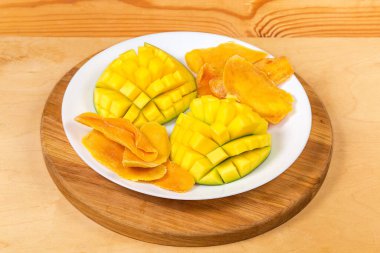 Dried slices of mango pulp and fresh mango halves cut in form the hedgehog style on white dish on a wooden surface
