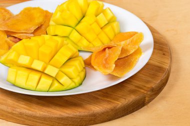 Dried slices of mango pulp and cut fresh mango halves on white dish on a wooden serving board, fragment close-up
