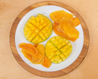 Dried slices of mango pulp and fresh mango halves cut in form the hedgehog style on white dish on a wooden surface, top view