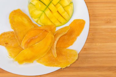 Dried slices of mango pulp and fresh mango half cut in form the hedgehog style on white dish on a wooden surface, fragment top view