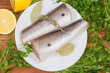 Kırsal bir masada taze maydanozun arasında beyaz bir tabakta baharatlı, başsız, çiğ Arjantin hake 'si.