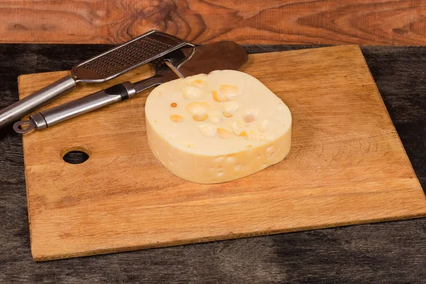 Piece of Swiss-type cheese on a wooden cutting board against the cheese slicer and cheese grater on dark table