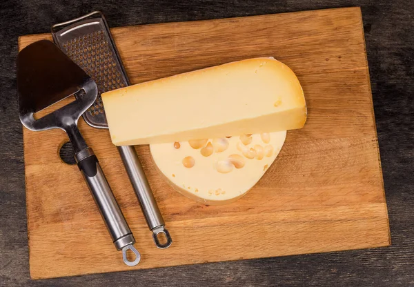 Pieces different shape of two types of semi-hard cheese, cheese slicer and cheese grater on a wooden cutting board, top view