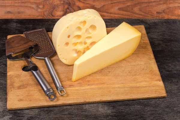 Pieces different shape of two types of semi-hard cheese, cheese slicer and cheese grater on a wooden cutting board