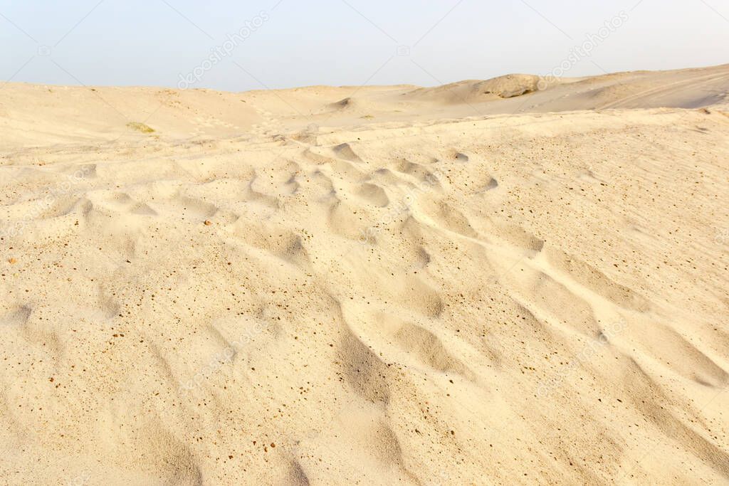 Slope of gray-yellow sandy dune with footprints sprinkled with windblown sand against clear sky