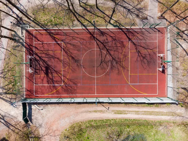 Universal outdoor sports field with red absorbing coating and color markings for playing various sports among the trees of the park in early spring, vertical aerial view