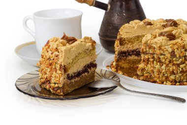 Kısmen katmanlı sünger keki, tereyağı kremalı, karamelize edilmiş süt, parçalanmış fındıklar küçük beyaz fincana karşı ve klasik bakır kahve demliğine karşı, beyaz arka planda bir parça.