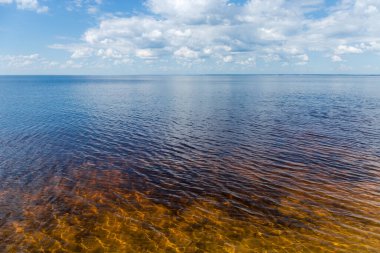 Hafif dalgalı su yüzeyi olan büyük bir su deposu. Yaz günlerinde hafif bulutlu, kumlu bir taban gökyüzüne karşı.