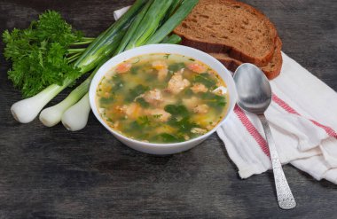 Beyaz kasede büyük balık kütleleri, peçetede kahverengi ekmek ve kaşık, koyu yüzeyde yeşil soğan ve maydanoz içeren somon çorbası.