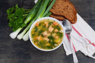 Beyaz kasede büyük balık kütleleri olan somon balığı çorbası, peçetede kahverengi ekmek ve kaşık, koyu yüzeyde yeşil soğan ve maydanoz.