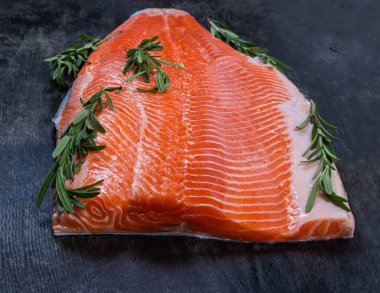 Taze biberiyenin dallarıyla birlikte teninde soğumuş çiğ gökkuşağı filetosu koyu bir yüzeye uzanıyor, seçici bir odak noktasında yakın plan.