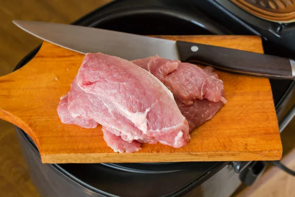 Pieces of uncooked pork pulp and kitchen knife lie on the wooden cutting board atop the open household multi-cooker before cooking, close-up in selective focus