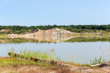 Terk edilmiş ilmenit madeninin bulunduğu yerde sığ bir göl var. Ormana karşı bir kıyıya dökülüyor ve yaz günü gökyüzü açık.