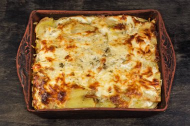 Kahverengi kabuklu patates güveci. Üstünde erimiş peynir var. Dikdörtgen kil fırın tepsisinde.