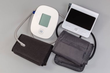 İki farklı elektronik sphygmomanometre, ölçüm birimleri ve şişirilebilir kelepçelerden oluşuyor. Gri arka planda kauçuk tüplerle bağlantılı, üst görünümde.