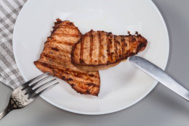 Çatal ve bıçaklı beyaz bir tabakta iki orta pişmiş hindi bifteği servis ederken, arka planda yakın plan gri bir görüntü.