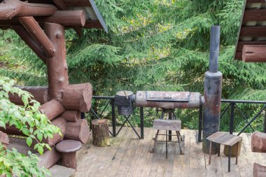 Metal fırın, barbekü ızgarası ve iki terası olan ahşap bir platformda açık ızgara alanı. Bulutlu bir günde geniş yapraklara karşı kalın kütükler ile inşa edilmiş.
