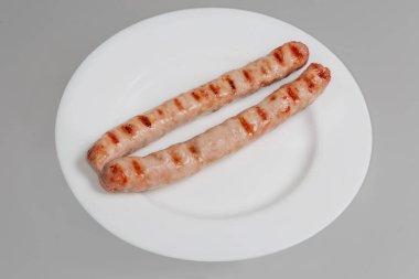 Izgara ızgara uzun domuz sosisi beyaz bir tabakta gri arka planda.