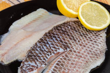 İki buzu çözülmüş çiğ dourade fileto derisi üzerinde kemiksiz balık leşi şeklinde siyah bir tabakta taze limon parçaları, yakın plan.