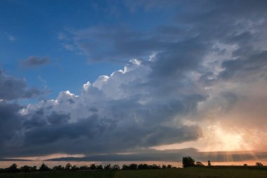 Mavi gökyüzünün kümülüs bulutları ve güneş ışınları olan bir bölümü gün batımında ağaçların tepesindeki bulutların ardından geliyor.