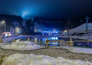 Ön planda bir kayak pistleri ve telesiyejler ve bir gölet ile ormanlık yamaca gece manzarası 