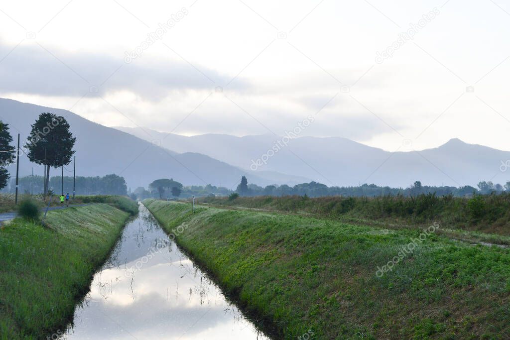 Hills with fog and clouds and river with water reflecting part of hills and clouds in the early hours of the morning and with two adults jogging