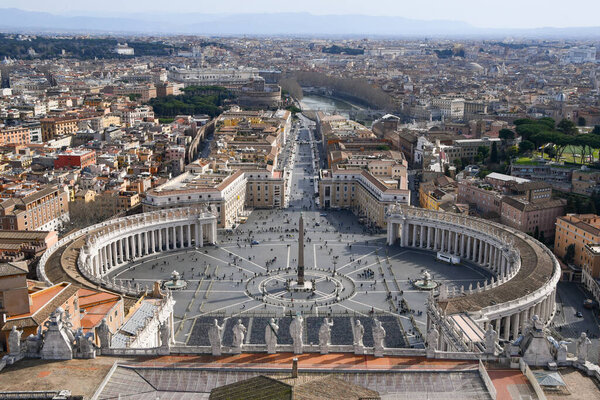 Overview from above on the elliptical arcade of St. Peter with roofs of nearby buildings and Via della Conciliazione and Tiber River with hills on the horizon and with few tourists around