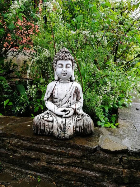 A garden statue of Buddha, a glazed ceramic Buddha figurine in a spring garden against a background of green leaves.  Mobile phone snapshot.  Illustration for Buddhist and yoga themes.