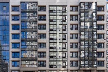 Bir grup mavi pencereli ve yansımalı balkonlu bej bir duvar. Modern bir binanın renkli pencerelerinin yatay dokusu