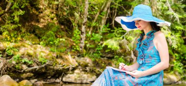 güzel genç kız nehirdeki bir kaya üzerinde oturur ve çizer