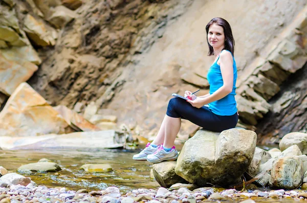 Bella ragazza siede su una roccia nel fiume e attinge da — Foto Stock