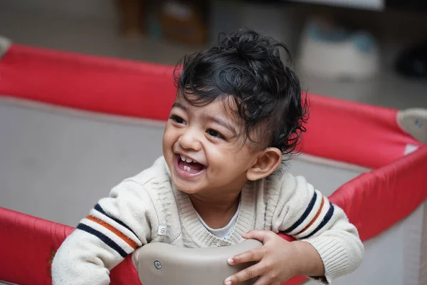 A closeup photo of an adorable indian toddler baby boy smiling with dimple in cheeks and standing inside a playpen — Stock Photo, Image