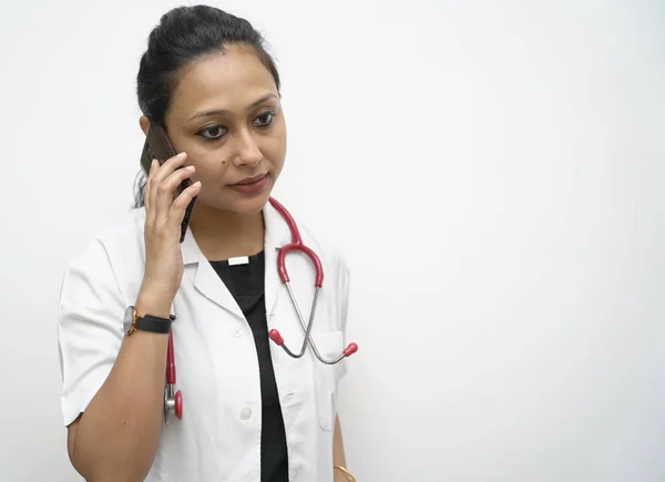 Uma médica indiana do sul nos anos 30 falando de telefone celular com casaco branco e estetoscópio vermelho em fundo branco. conceito de telemedicina e teleconsulta — Fotografia de Stock