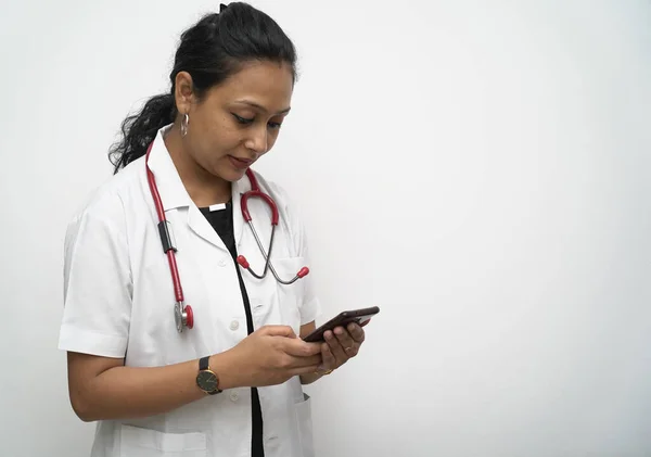 Індійська жінка-лікар у 30-х роках перевіряє мобільний телефон у білому пальто та червоний стетоскоп на білому фоні. — стокове фото