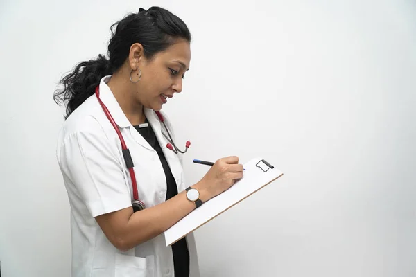 Южноиндийская женщина-врач 30 лет выписывает медицинские рецепты на доске с ручкой в белом халате и красным стетоскопом — стоковое фото